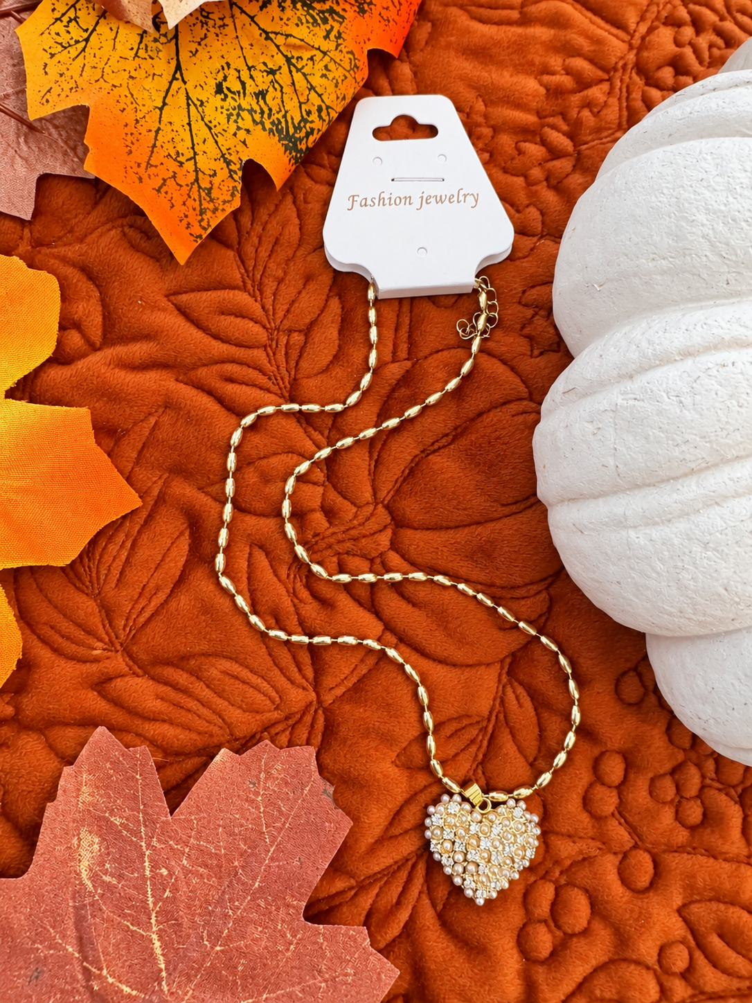 A gold heart-shaped necklace with clear rhinestones on a gold chain, attached to a white card labeled 'Fashion jewelry' and displayed on an orange quilted blanket with autumn leaves and a white pumpkin.