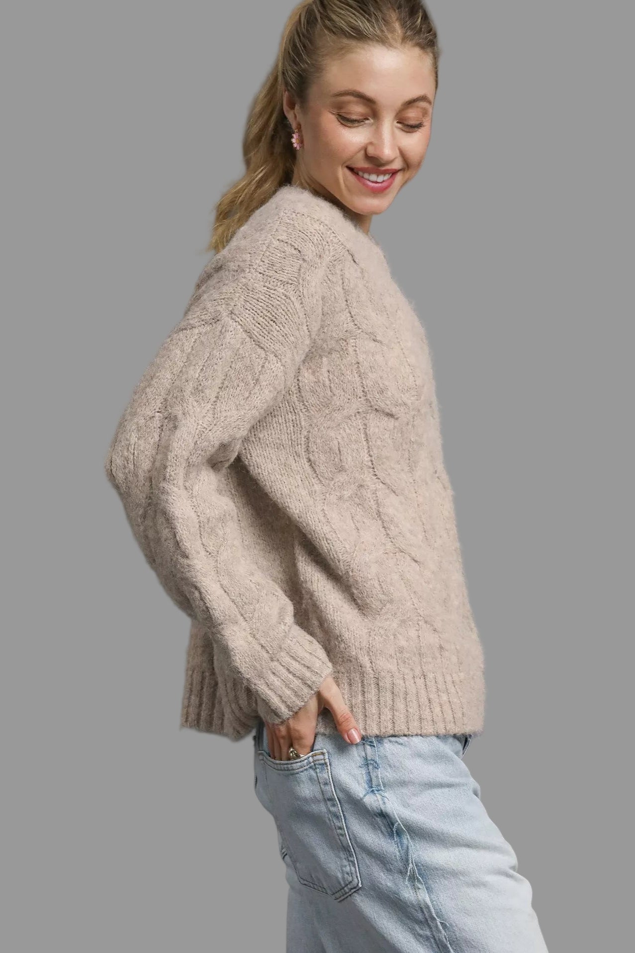 Woman wearing a beige knitted sweater and light blue jeans against a gray background
