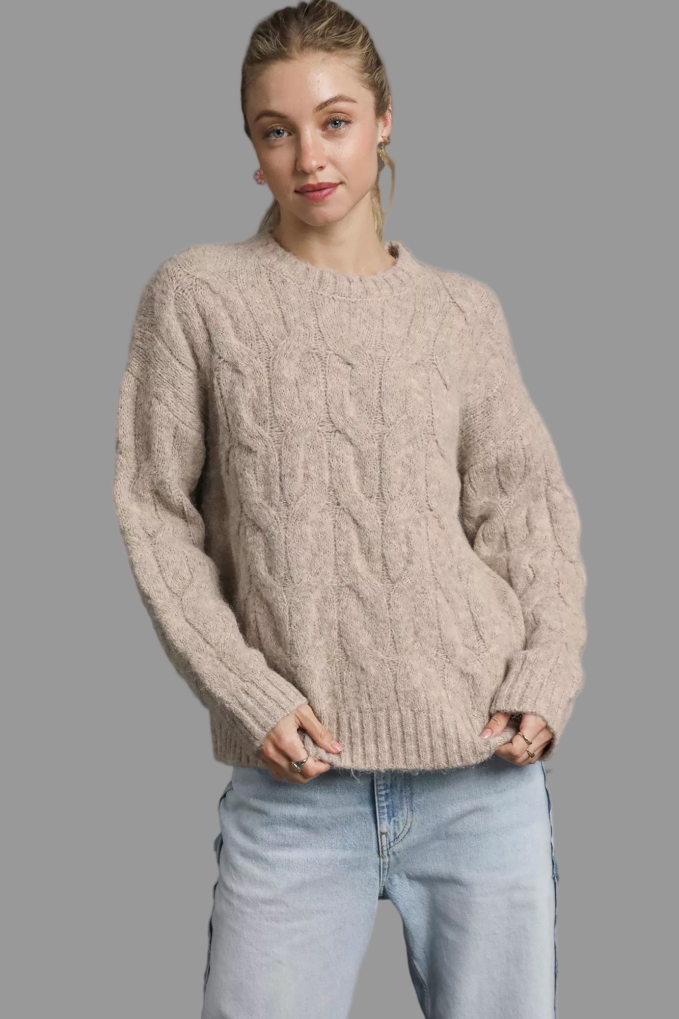 Woman wearing a beige cable knit sweater and light blue jeans on a white background