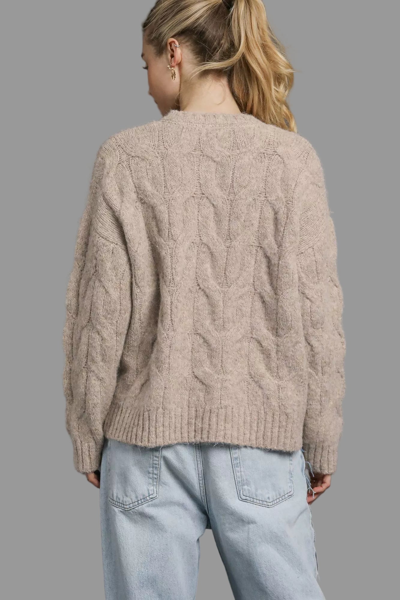 Person wearing a beige cable knit sweater and light blue jeans on a gray background