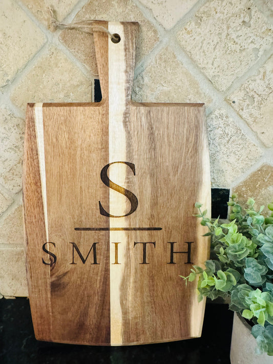 Wooden cutting board with engraved initials and name, placed on a tiled wall.