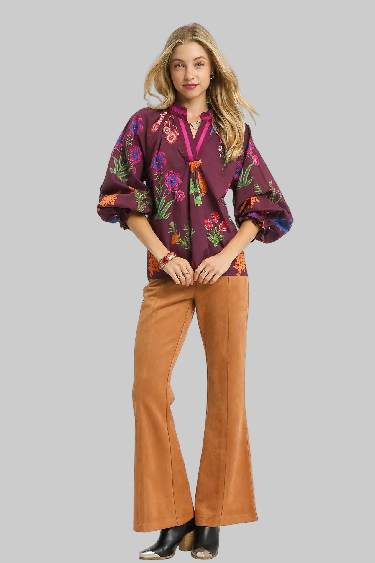 Woman wearing a burgundy floral blouse and tan bell-bottom pants on a white background