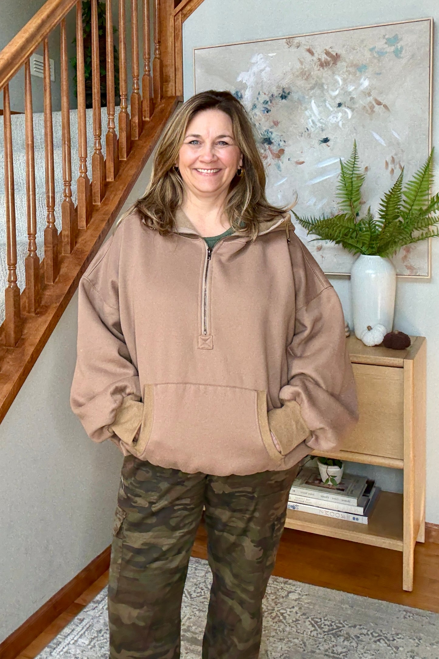 Woman wearing a brown hoodie 1/2 zip sweatshit and camouflage pants standing in a home setting with a staircase and decor.