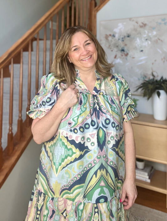 Woman wearing a colorful patterned dress standing in a home setting with a staircase and decor.