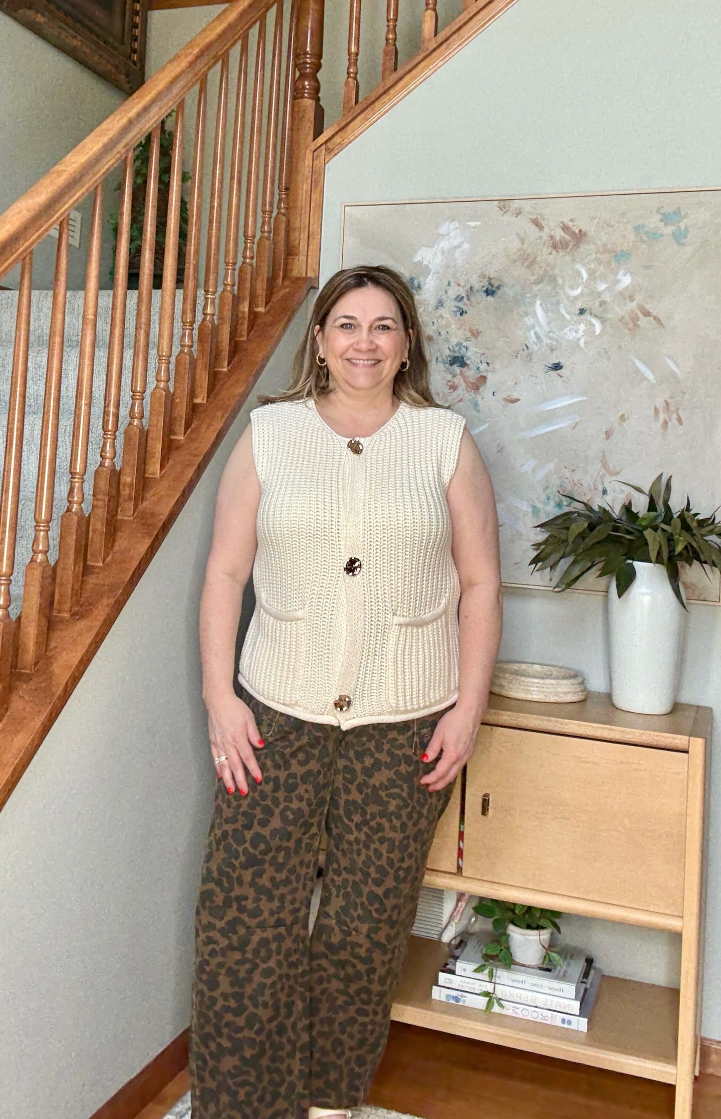 Woman standing in a home setting with wooden staircase and bookshelf.