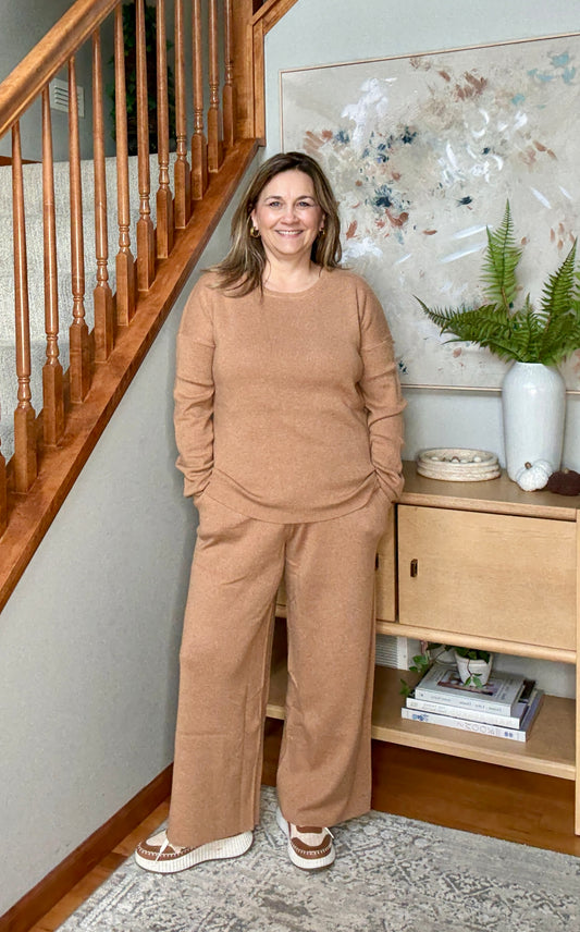 Woman in a beige outfit standing in a home setting with a staircase and decorative items.