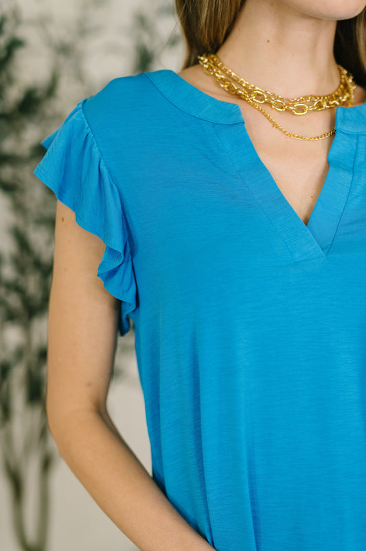 Person wearing a blue top with ruffled sleeves and gold necklaces against a blurred background