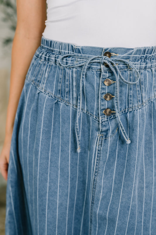 Close-up of a person wearing a blue denim skirt with thin white vertical pinstripes, an elastic drawstring waist, and front buttons, paired with a white top.
