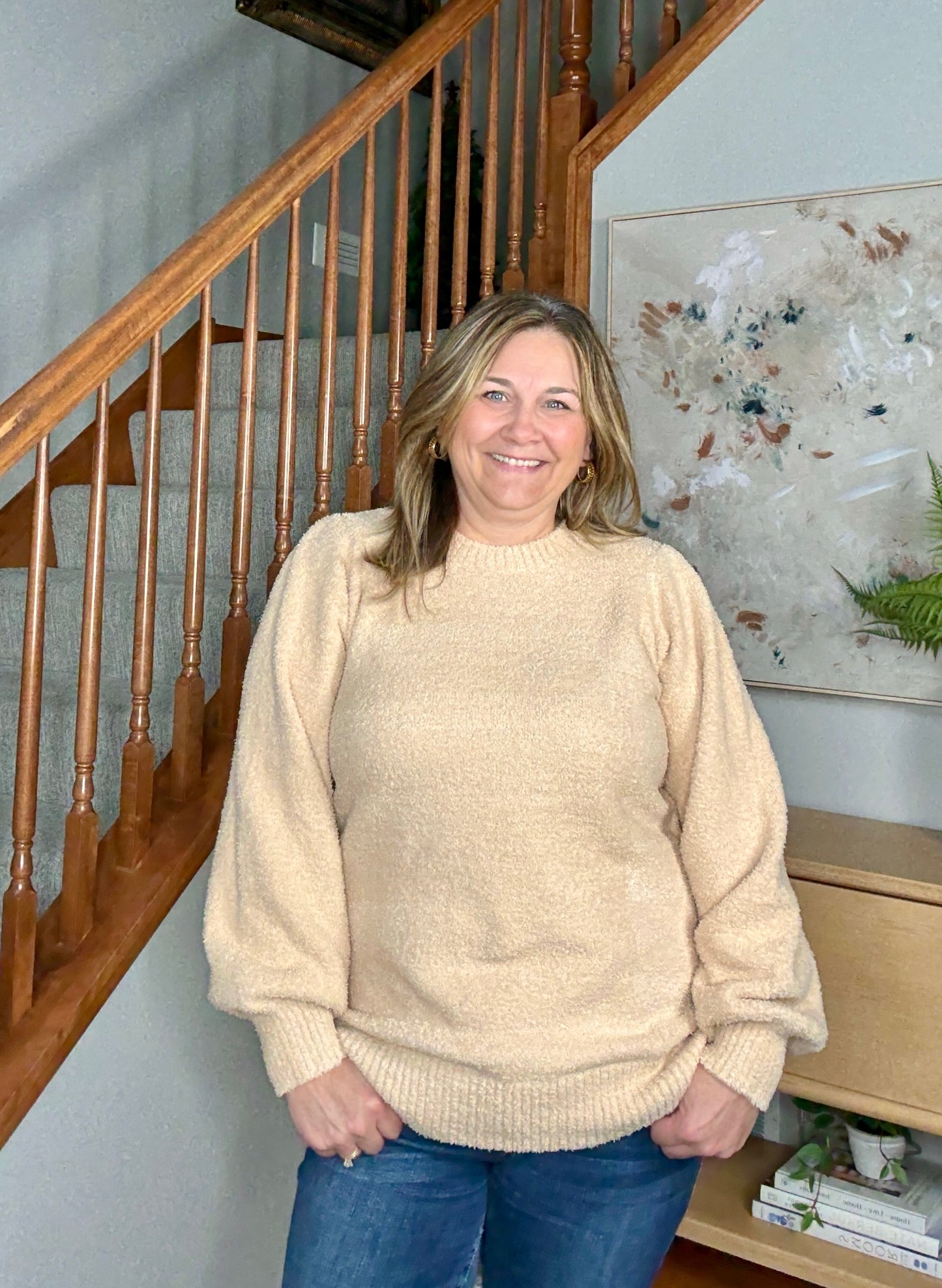 Woman wearing a beige sweater standing in a home setting with a staircase and artwork in the background.