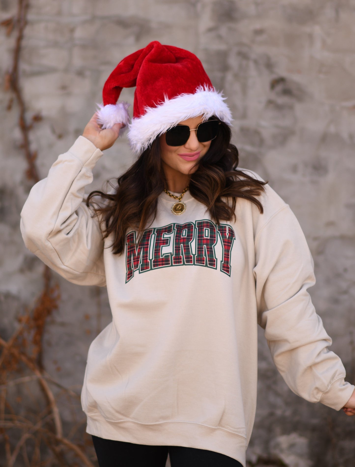 Person wearing a beige sweatshirt with 'MERRY' text, a red Santa hat, and sunglasses against a stone wall.