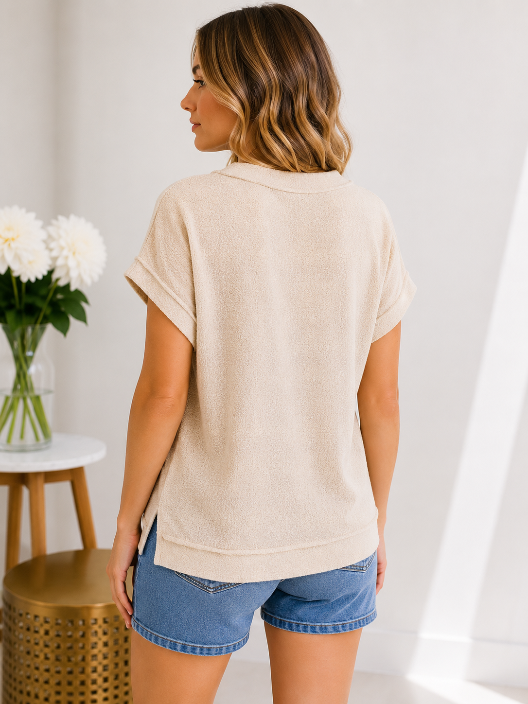 Woman wearing a beige short-sleeve shirt and denim shorts in a minimal indoor setting.