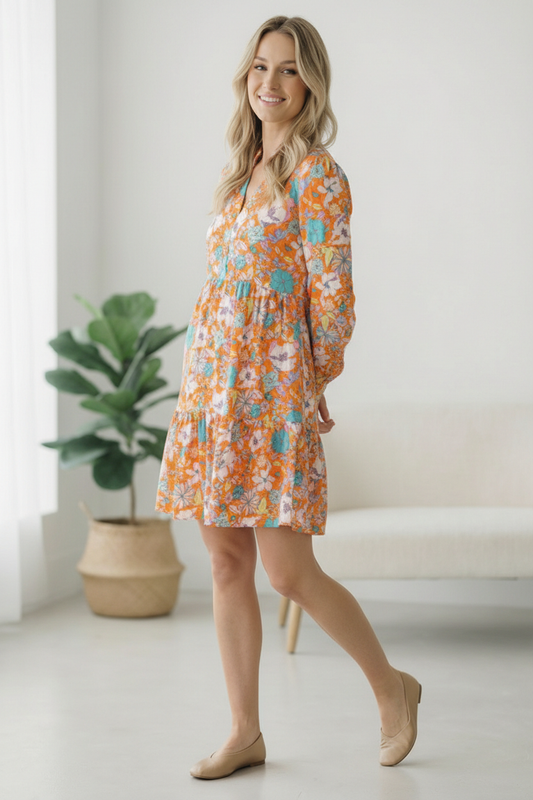 Woman wearing an orange floral dress standing in a bright room with a plant and white sofa.