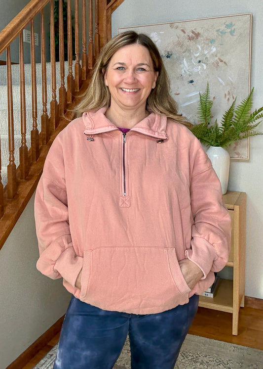 Woman wearing a pink pullover in a home setting with a staircase and plant in the background