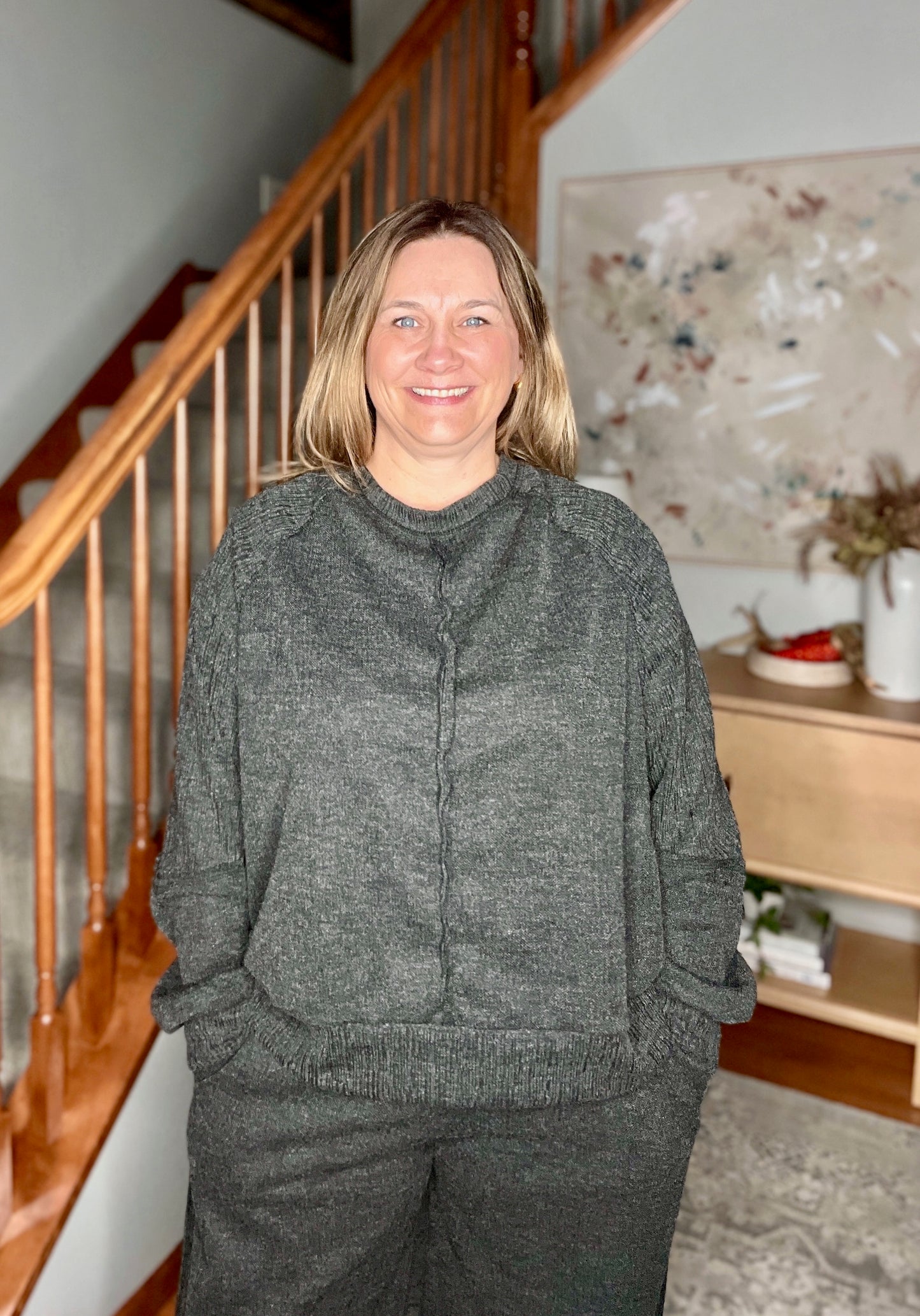 Woman wearing a gray sweater and pants standing in a home setting with a staircase and decorative items in the background.