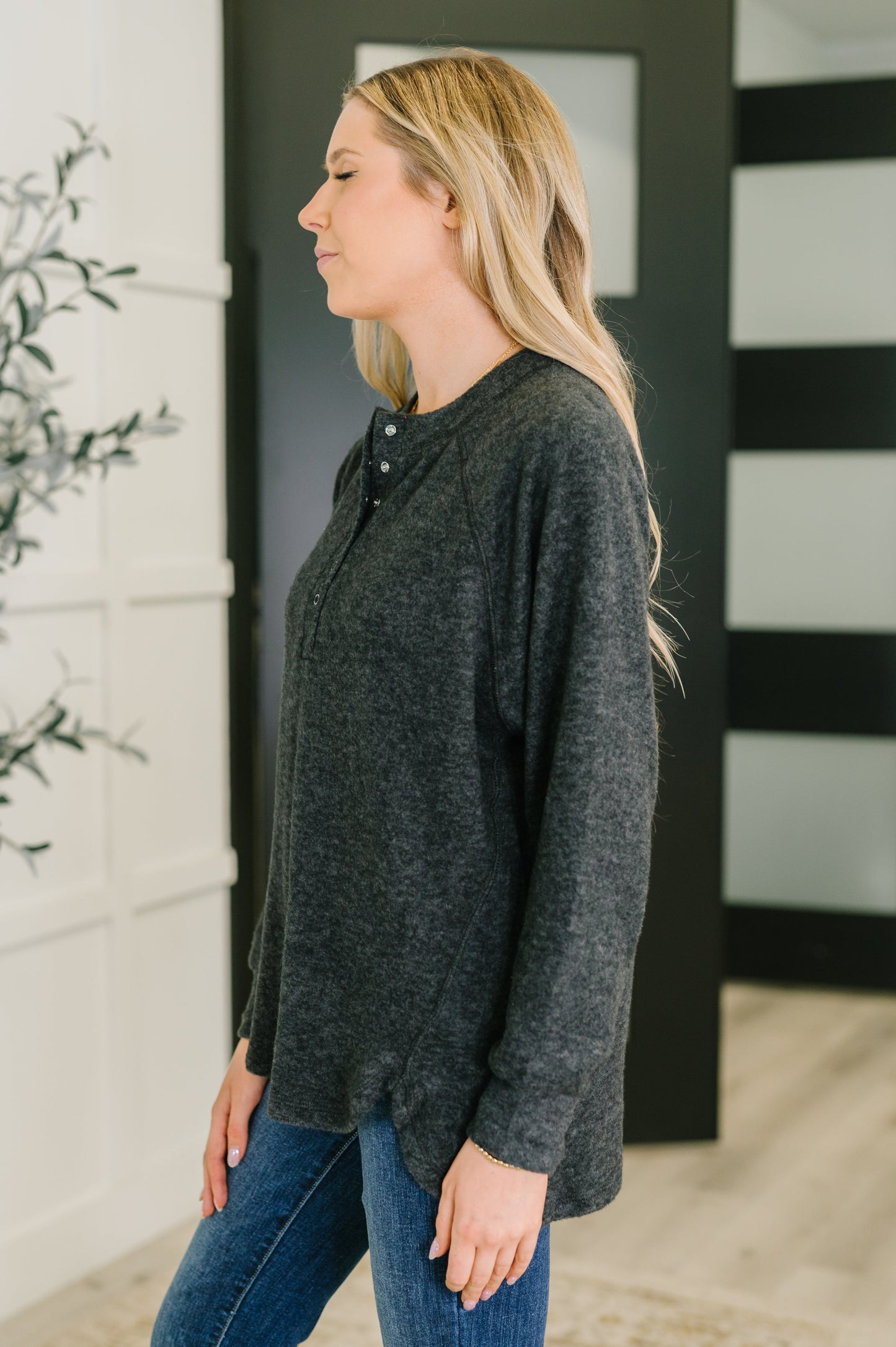 Woman wearing a black hi lo henley long-sleeve top and blue jeans standing against a black and white striped wall.