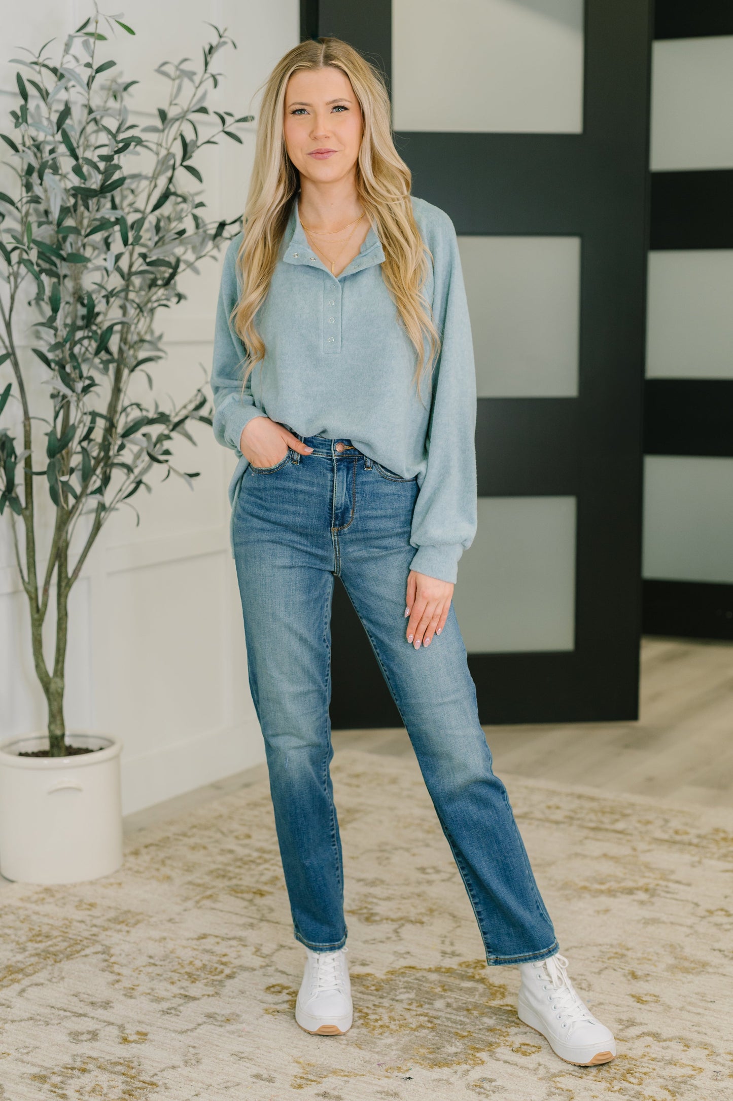 Woman wearing a light blue snap henley long sleeve shirt and jeans standing indoors with a plant and decorative wall in the background.