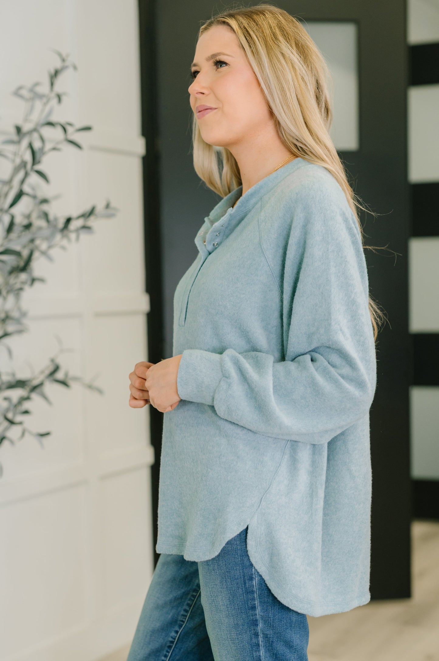 Woman wearing a light blue hi lo henley sweater and jeans standing indoors.