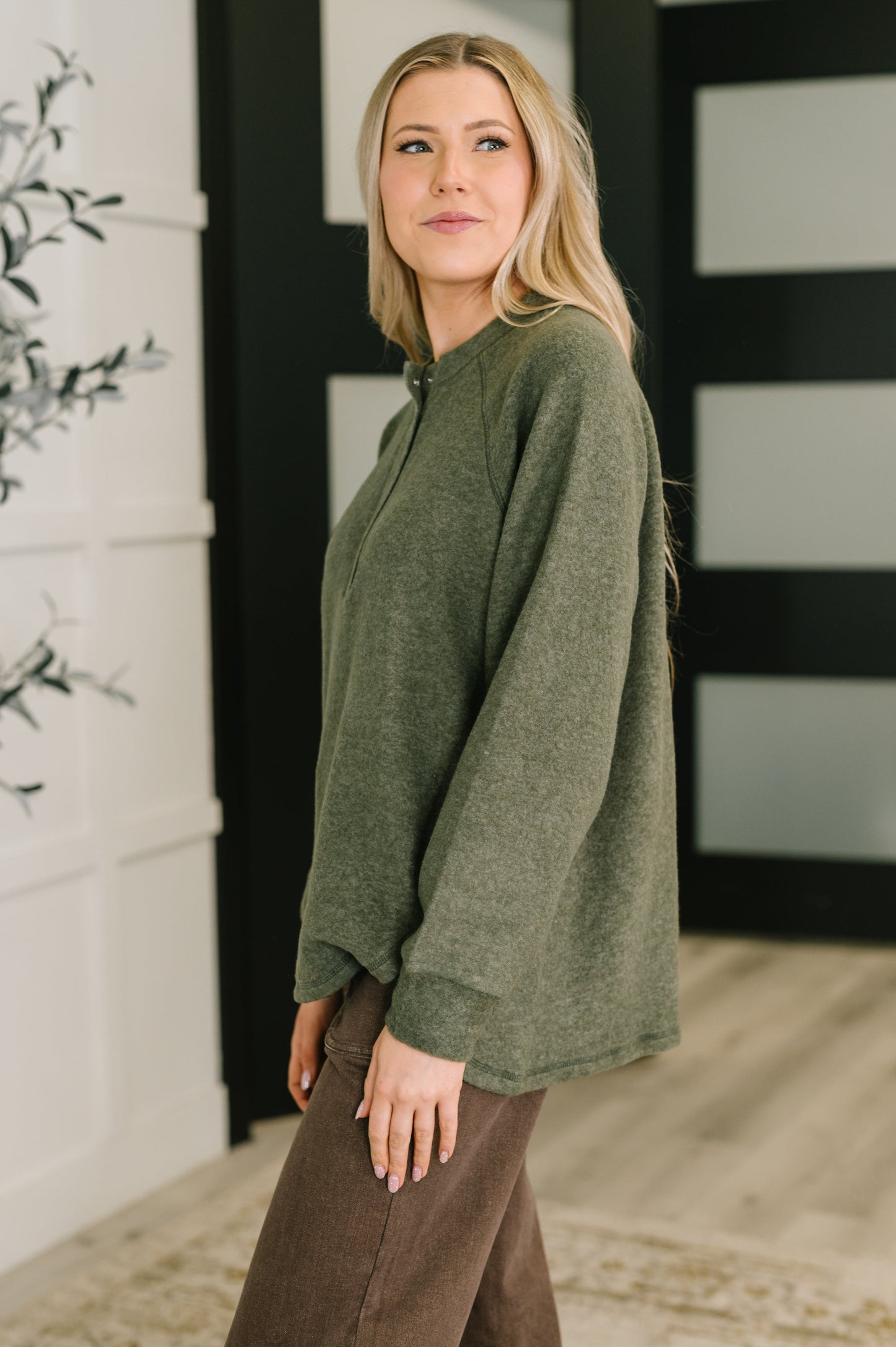 Woman wearing an olive green snap henley sweater and brown pants standing in a room with decorative elements.