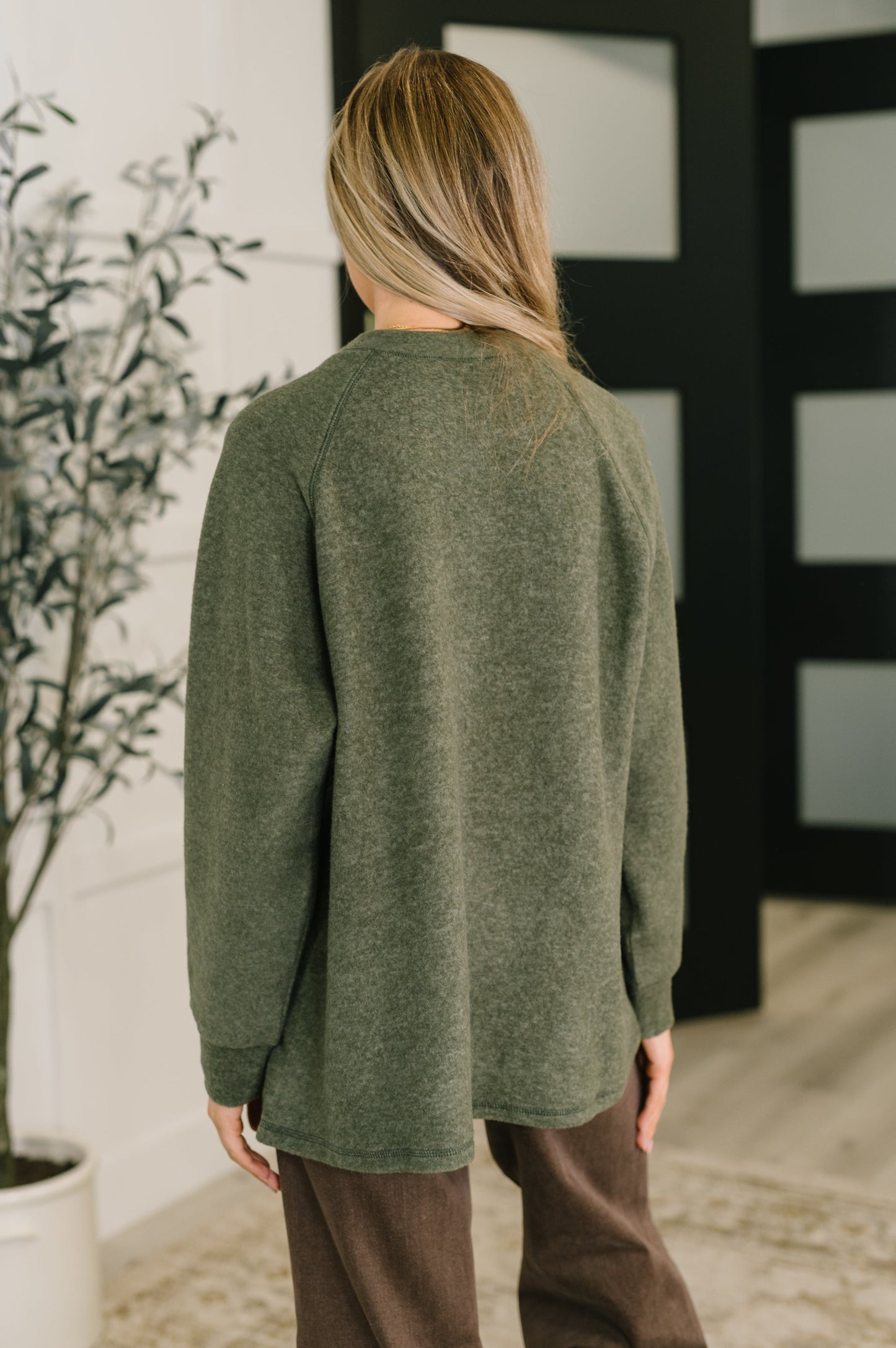 Person wearing an olive henley sweater and brown pants standing indoors with a plant and wall decor in the background.