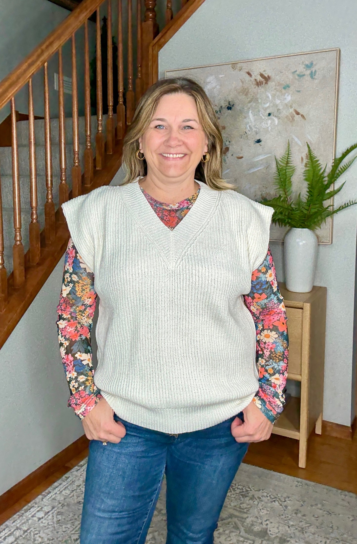 Woman wearing a cream sweater vest over a colorful shirt with floral sleeves, standing in a home setting.
