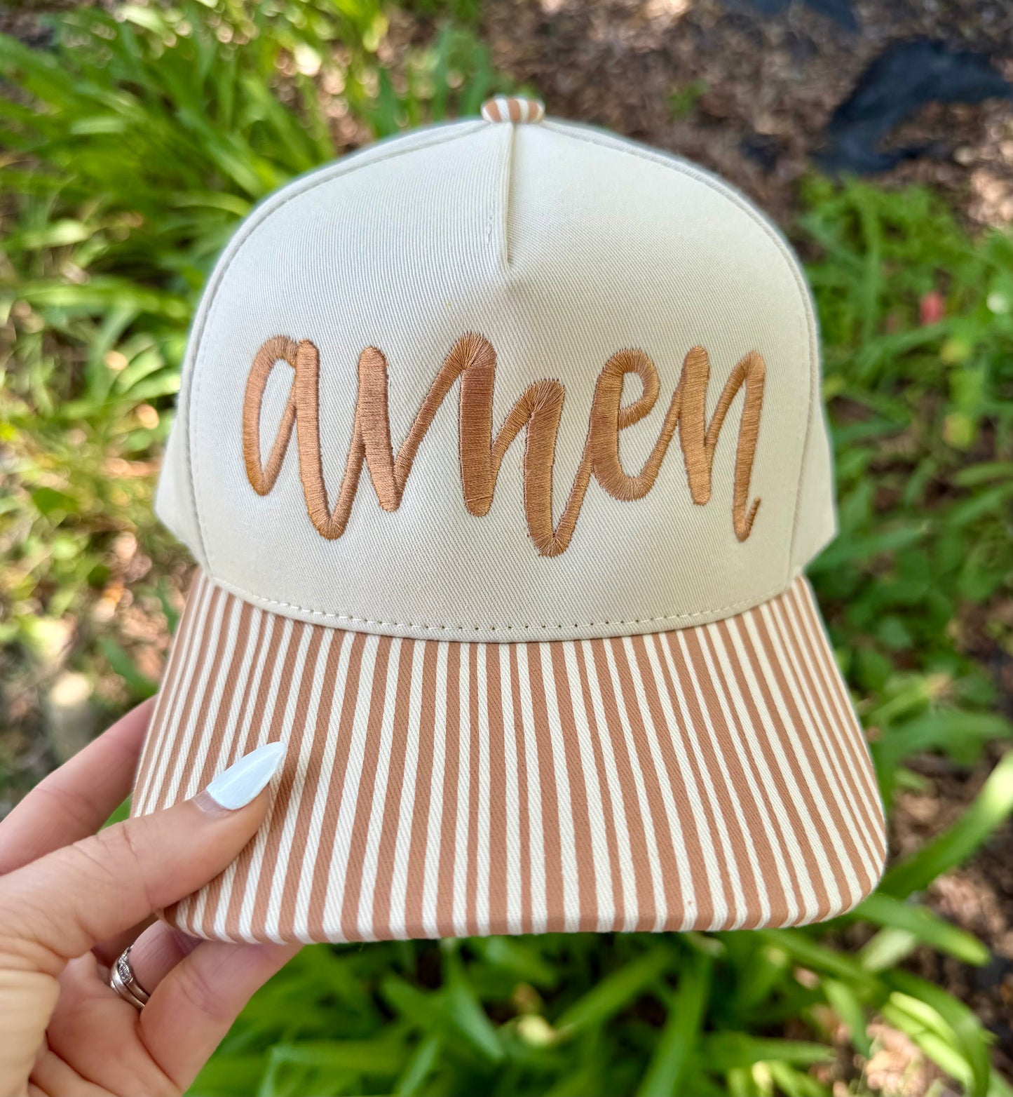 A hand holds a baseball cap outdoors with green foliage in the background. The cap has an off-white crown with metallic copper embroidered script reading 'amen' and a matching off-white and copper striped visor.