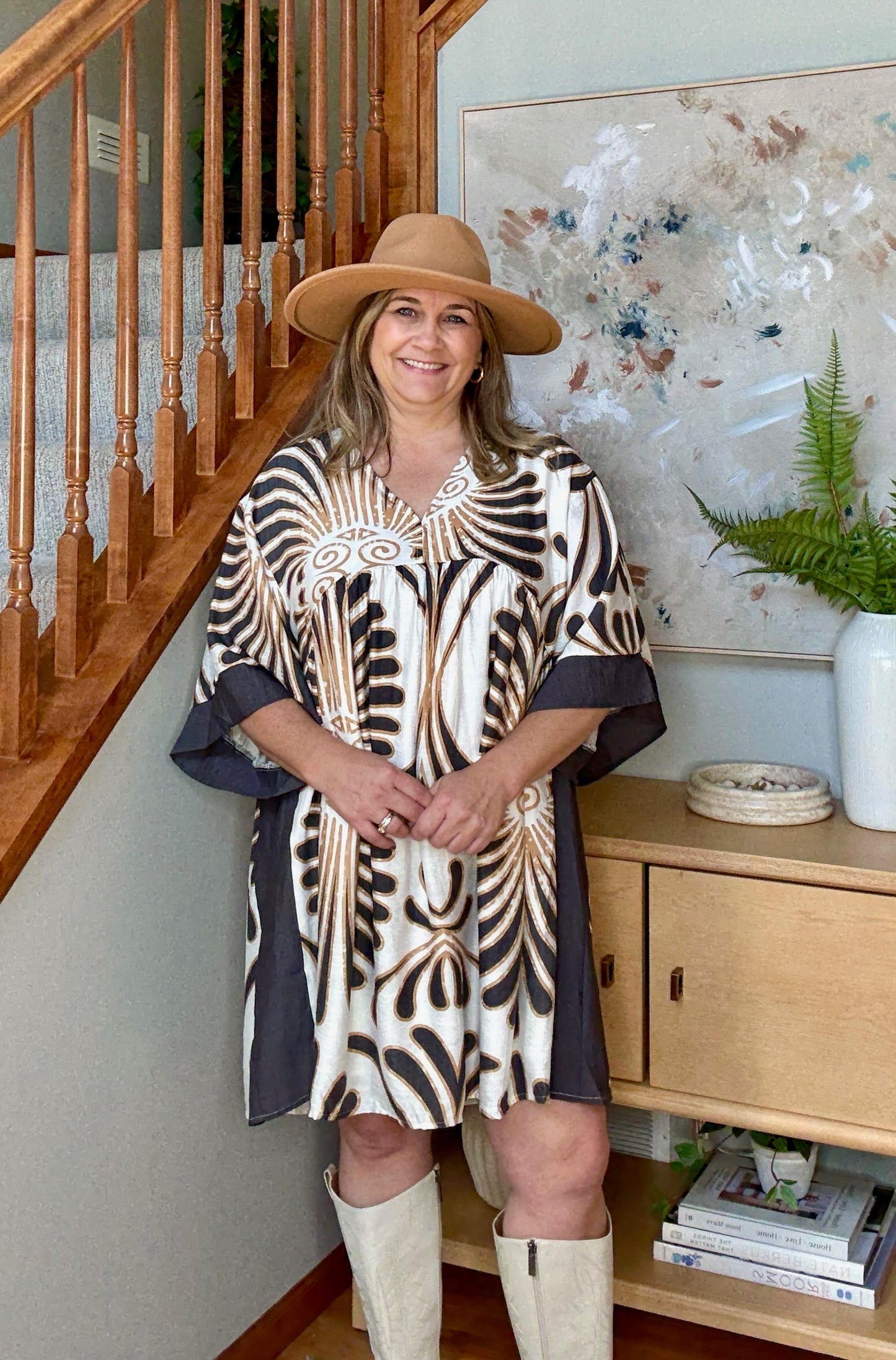 Woman in a patterned print dress and wide-brimmed hat standing in a home interior.