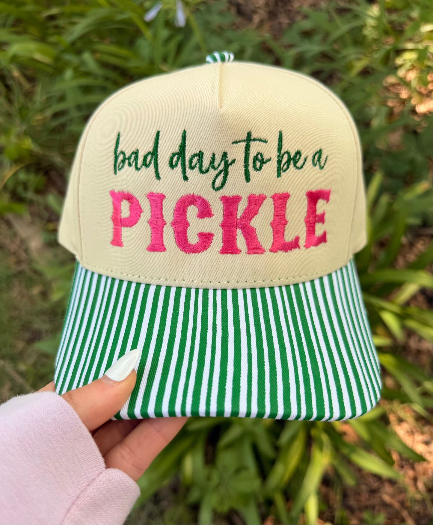 Cap with 'bad day to be a pickle' text held by a hand against a green outdoor background