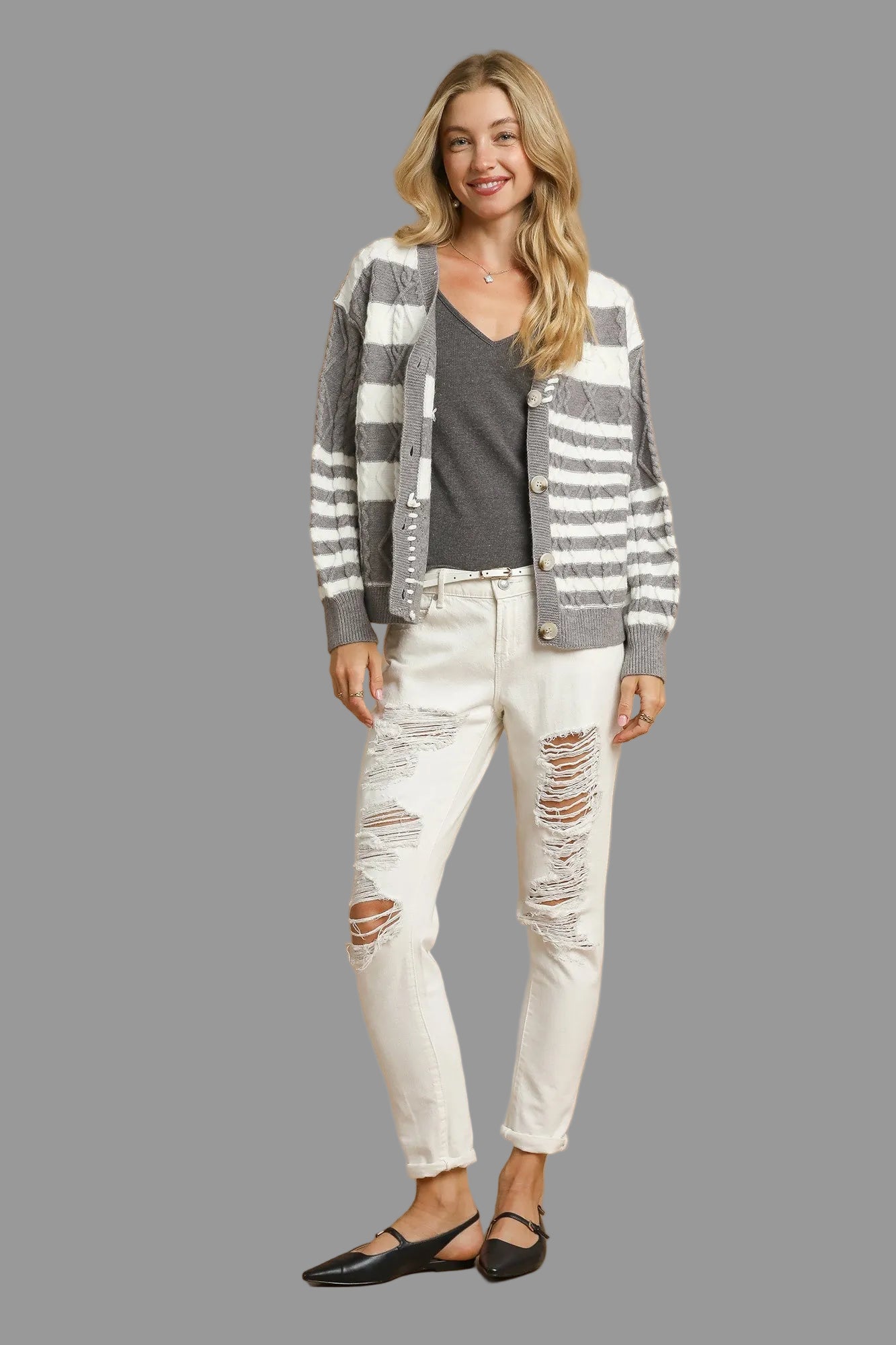 Woman wearing a striped button up cardigan, grey top, and white distressed jeans on a grey background