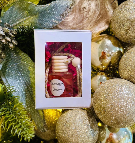Small bottle of cinnamon vanilla scented car diffuser in a decorative box surrounded by gold Christmas ornaments.