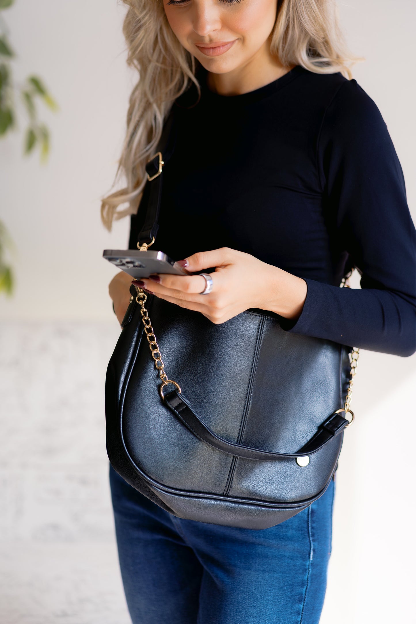 Woman holding a black handbag and looking at her phone against a light background