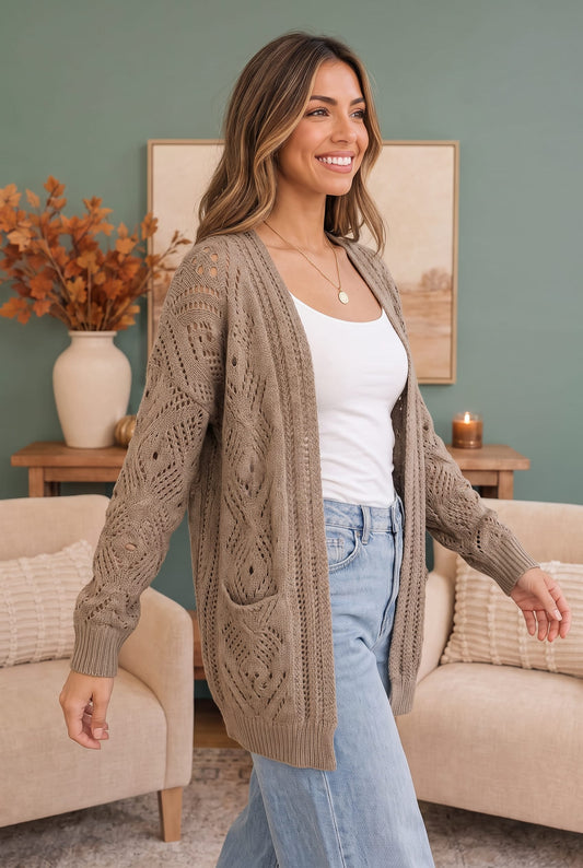 A woman wearing a taupe cable-knit cardigan over a white tank top and light blue jeans, standing in a cozy living room setting with beige sofas and a green wall.