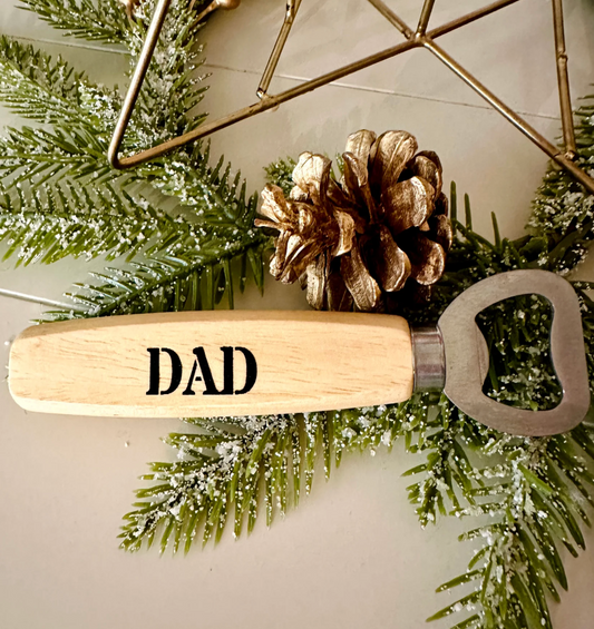 Bottle opener with 'DAD' engraving on a decorative wreath with pine cones and greenery.