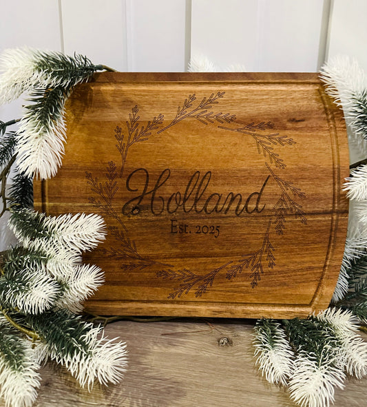 Wooden cutting board with engraved 'Holland Est. 2023' surrounded by decorative greenery.