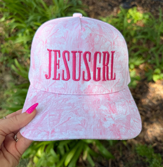 A pink and white marbled baseball cap with 'JESUSGRL' embroidered in pink on the front, held by a hand with bright pink fingernails against a blurred green garden background.