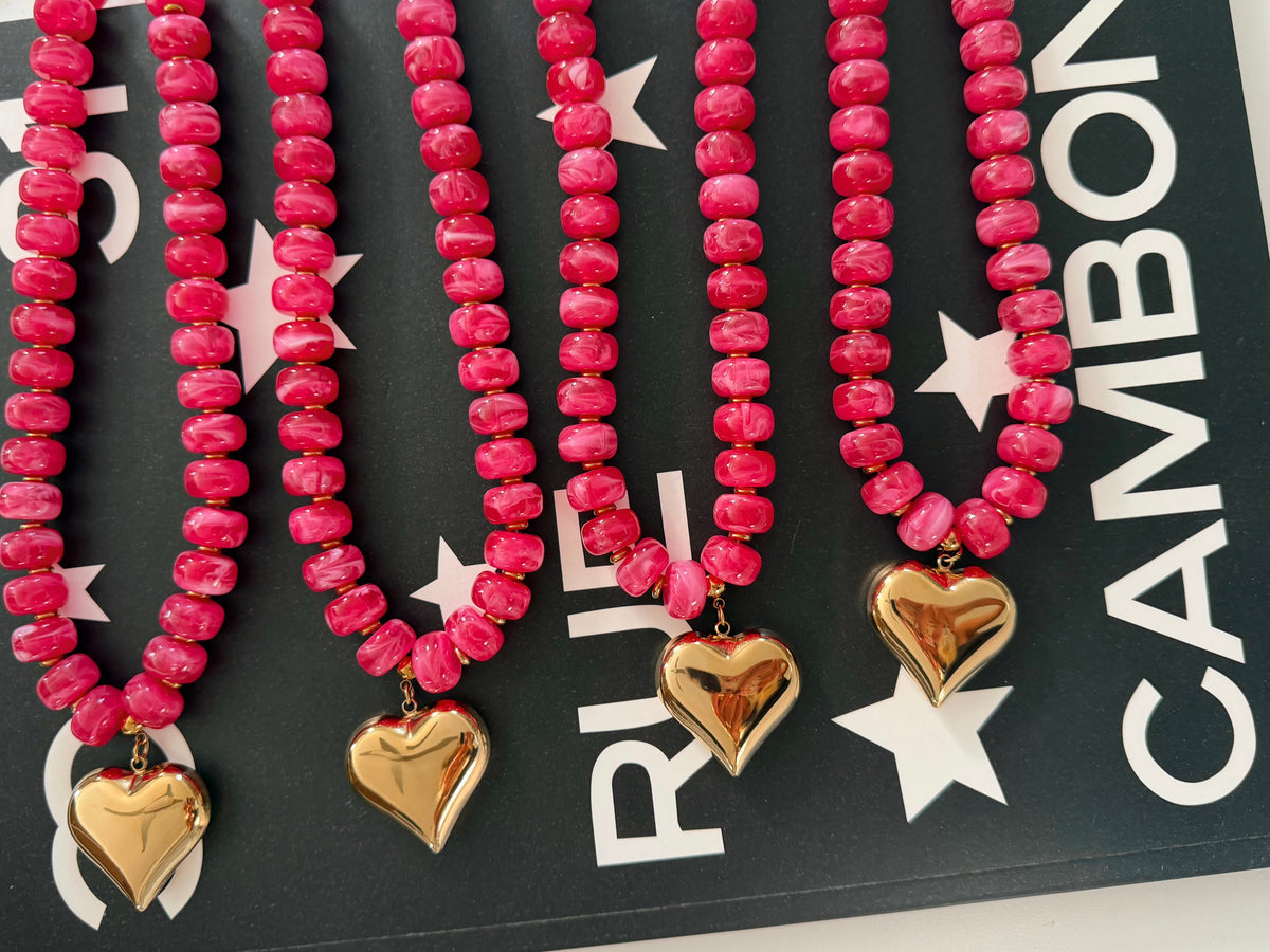 Four pink beaded necklaces with gold heart charms arranged diagonally on a black card with white text and star graphics.