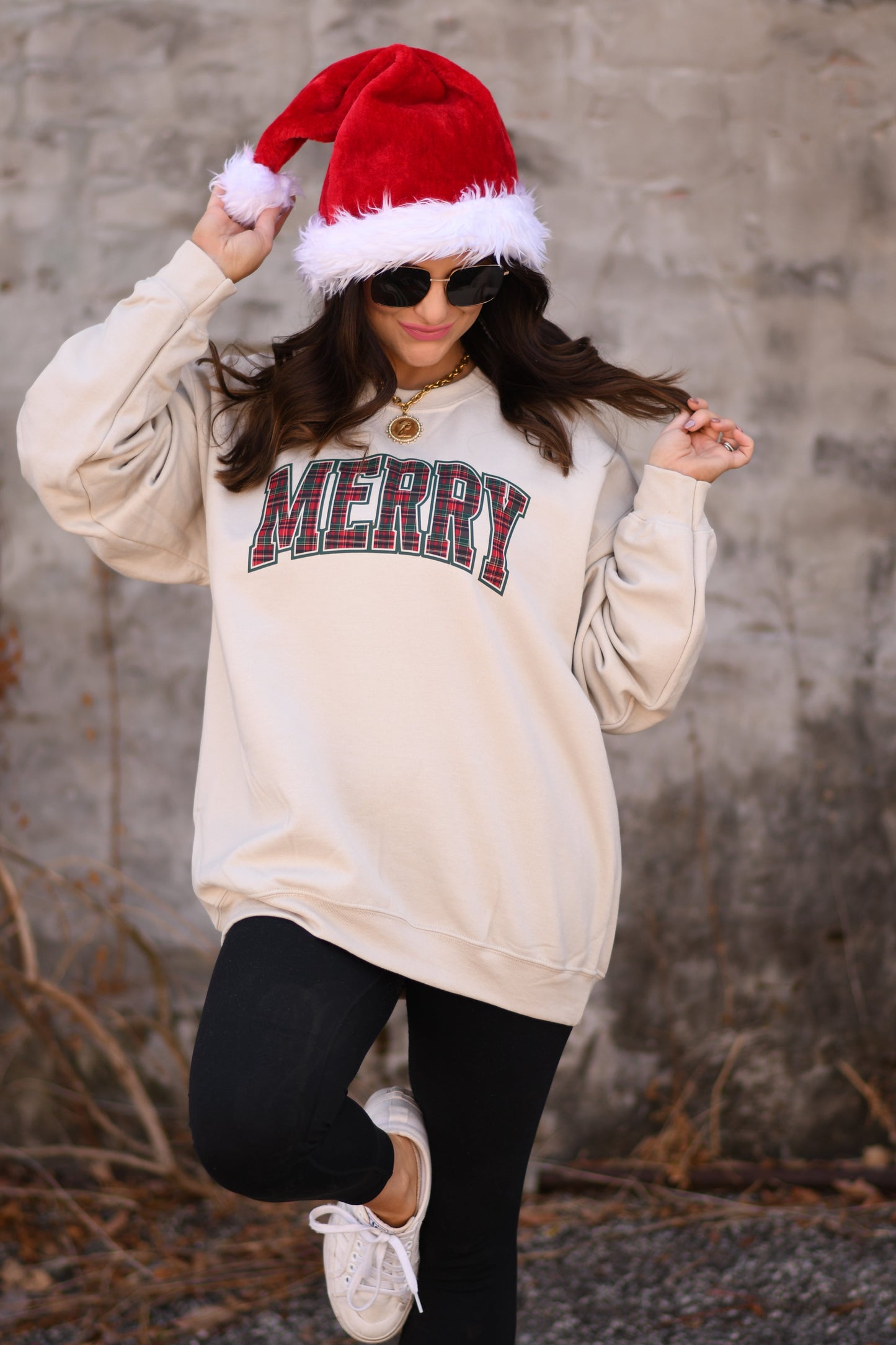Person wearing a beige sweatshirt with 'MERRY' text, black leggings, white sneakers, and a red Santa hat against a gray wall.
