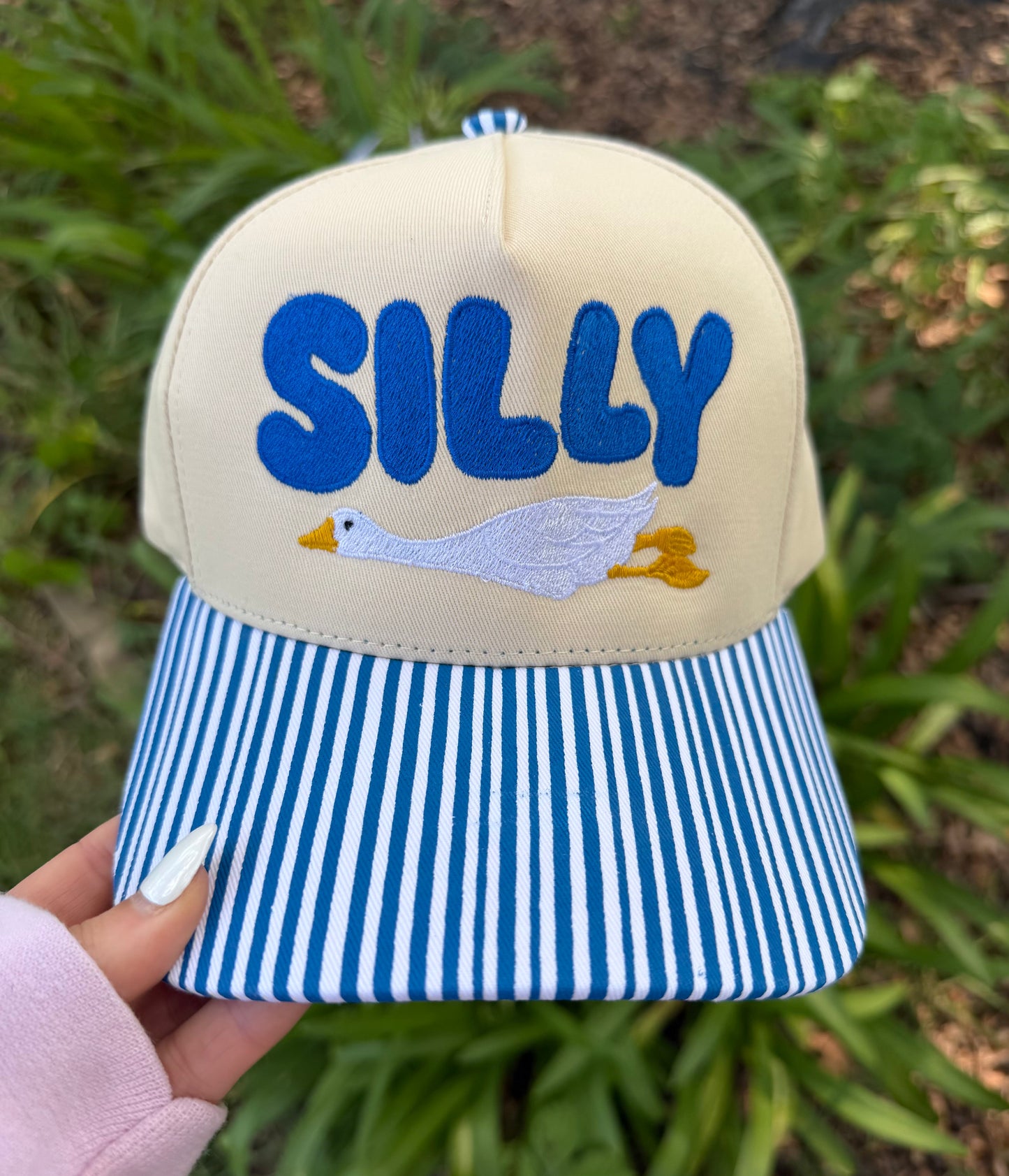 Beige cap with blue 'SILLY' text and duck design, held against a grassy background.