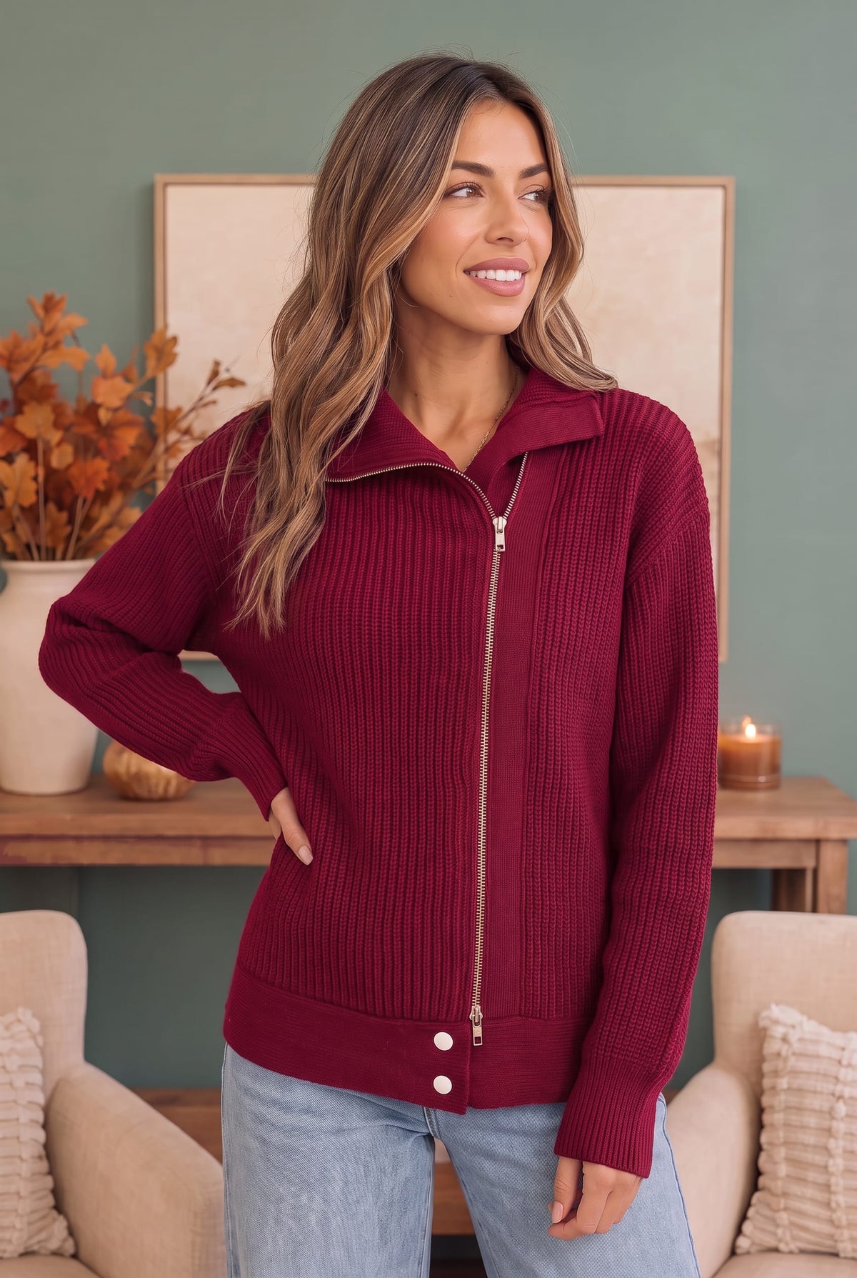 A woman wearing a ribbed knit zip-up sweater in deep burgundy with a stand collar and front zipper, paired with light blue jeans, standing in a cozy living room setting.