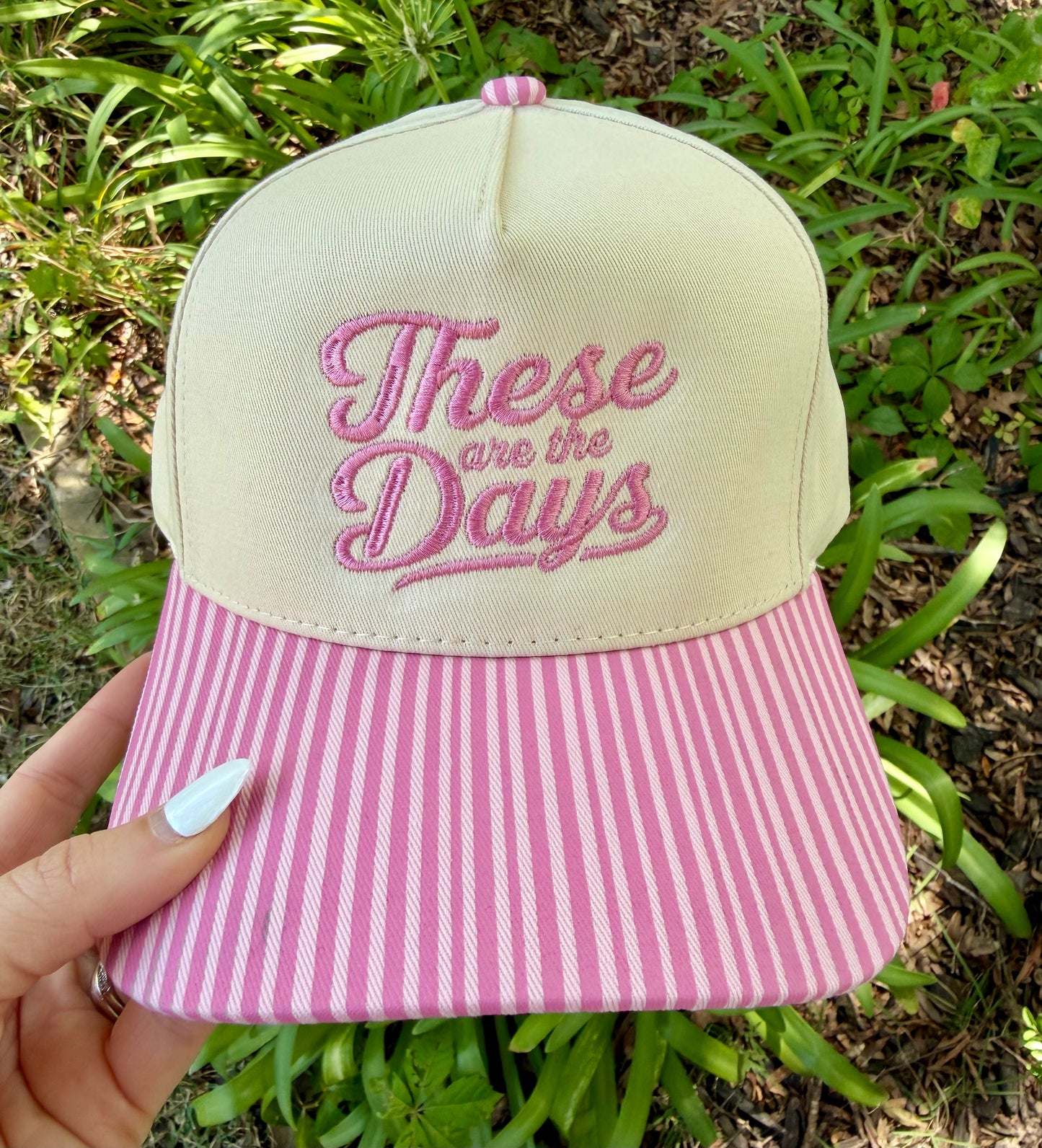 A hand holds a baseball cap outdoors with green foliage in the background. The cap has a cream crown with pink embroidered text reading 'These are the Days' and a pink-and-white vertically striped brim.