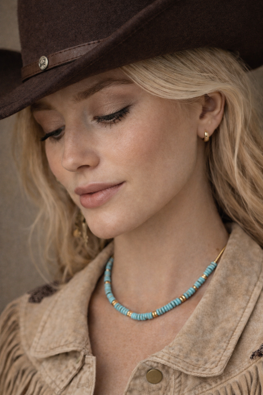 Woman wearing a brown cowboy hat and beige jacket with a blue beaded necklace.