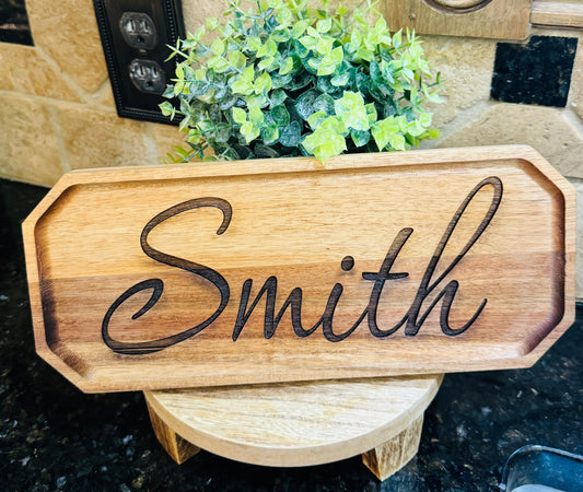 Wooden sign with 'Smith' engraved on it, placed on a surface with a plant in the background.