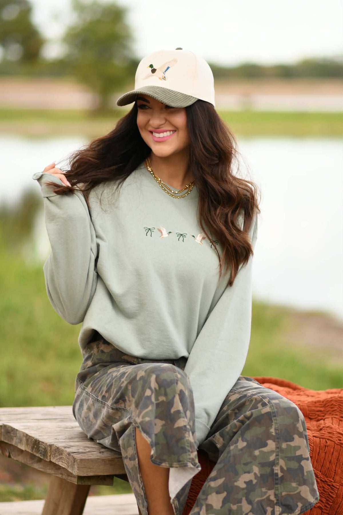 A woman sitting on a wooden bench outdoors by a body of water, wearing a sage green crewneck sweatshirt with embroidered birds, camouflage pants, and a beige baseball cap.