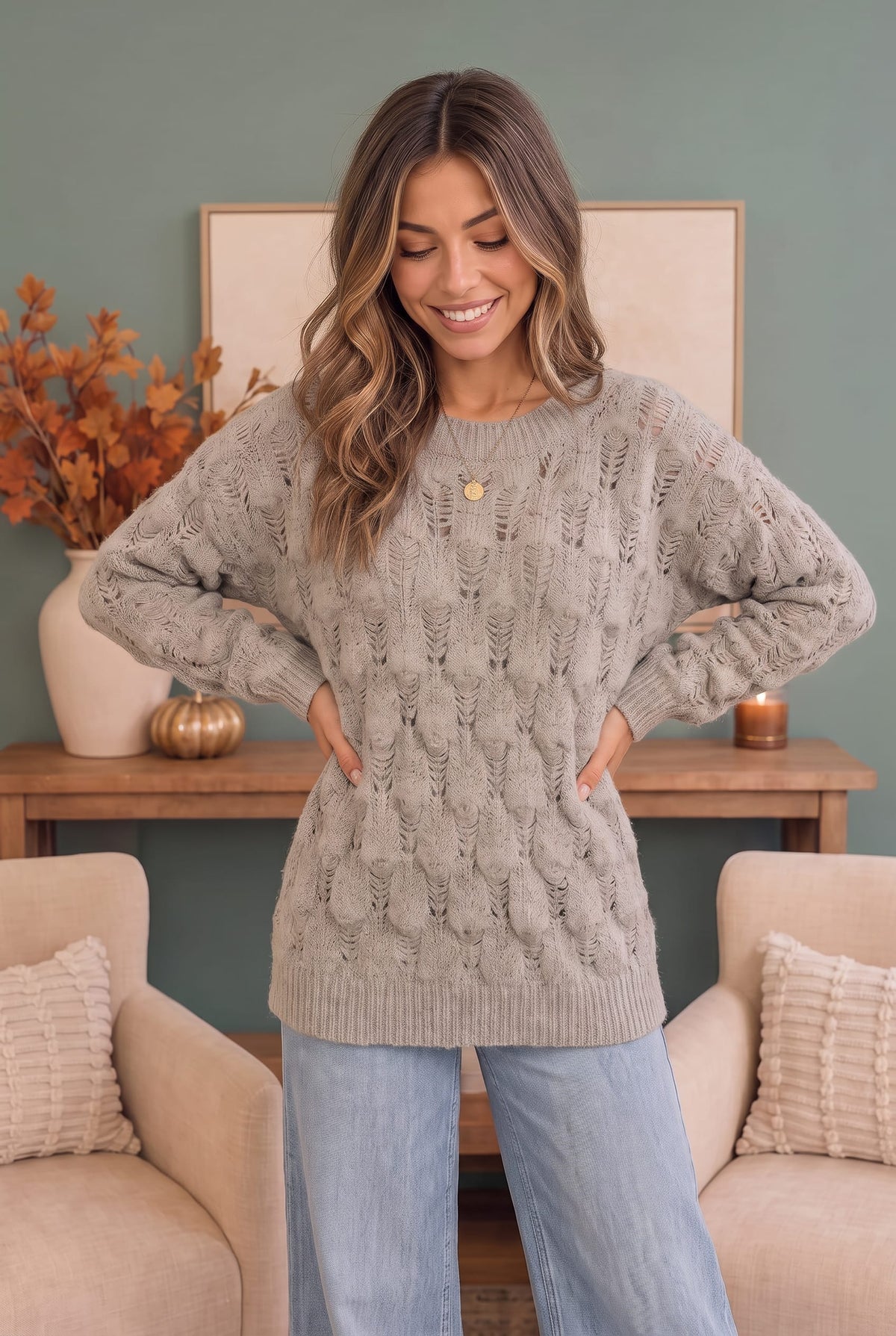 A woman wearing a chunky cable-knit crewneck sweater in taupe gray, standing in a cozy living room setting with beige armchairs and a wooden console table.
