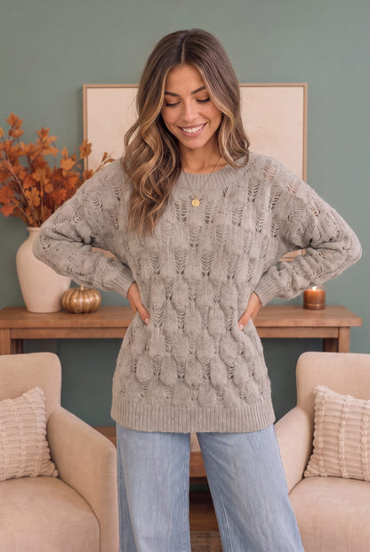 A woman wearing a chunky cable-knit crewneck sweater in taupe gray, standing in a cozy living room setting with beige armchairs and a wooden console table.