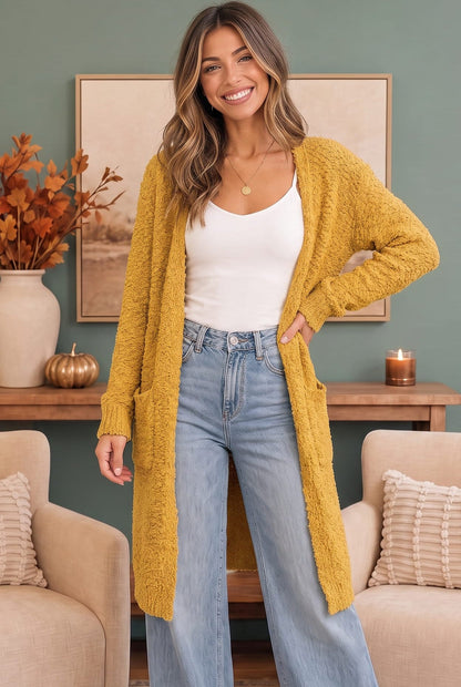 A woman stands in a living room wearing a long, fuzzy mustard yellow cardigan over a white tank top and light blue jeans.