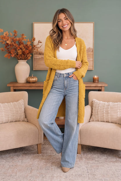 A woman stands in a living room wearing a mustard yellow knit cardigan over a white tank top and light blue wide-leg jeans. The room has a sage green wall, a wooden console table with a vase of dried leaves, and two beige armchairs.