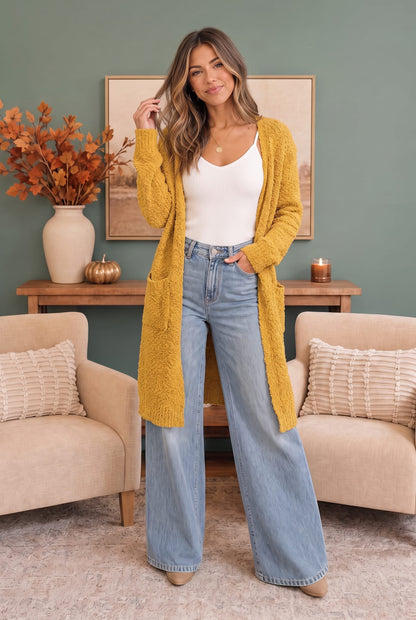A woman stands in a living room wearing a mustard yellow fuzzy open-front cardigan over a white tank top and light blue wide-leg jeans.