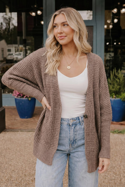 Woman wearing a brown cardigan, white top, and blue jeans outdoors.