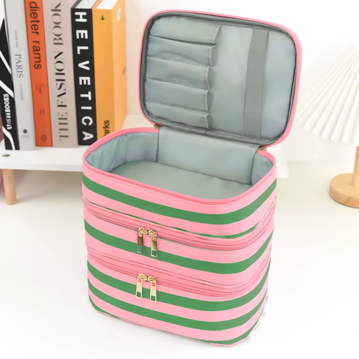 A pink and green striped cosmetic organizer case with an open lid showing interior compartments, photographed on a white tabletop with books and a lamp in the background.