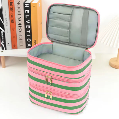A pink and green striped cosmetic organizer case with an open lid showing interior compartments, photographed on a white tabletop with books and a lamp in the background.